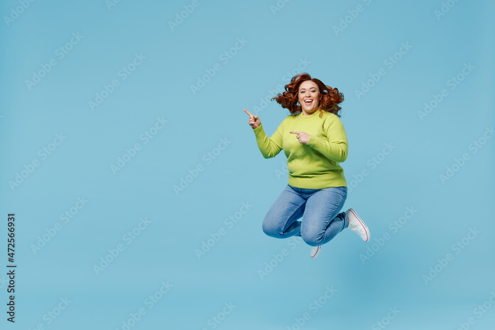 Young overjoyed smiling chubby overweight plus size big fat fit woman wear green sweater jump high point index finger aside on copy space isolated on plain blue background. People lifestyle concept