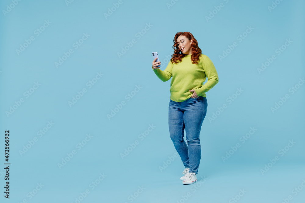 Full body smiling fun young chubby overweight plus size big fat fit woman wearing green sweater hold use mobile cell phone isolated on plain blue background studio portrait. People lifestyle concept. Full body smiling fun young chubby overweight plus size big fat fit woman wearing green sweater hold use mobile cell phone isolated on plain blue background studio portrait. People lifestyle concept.
