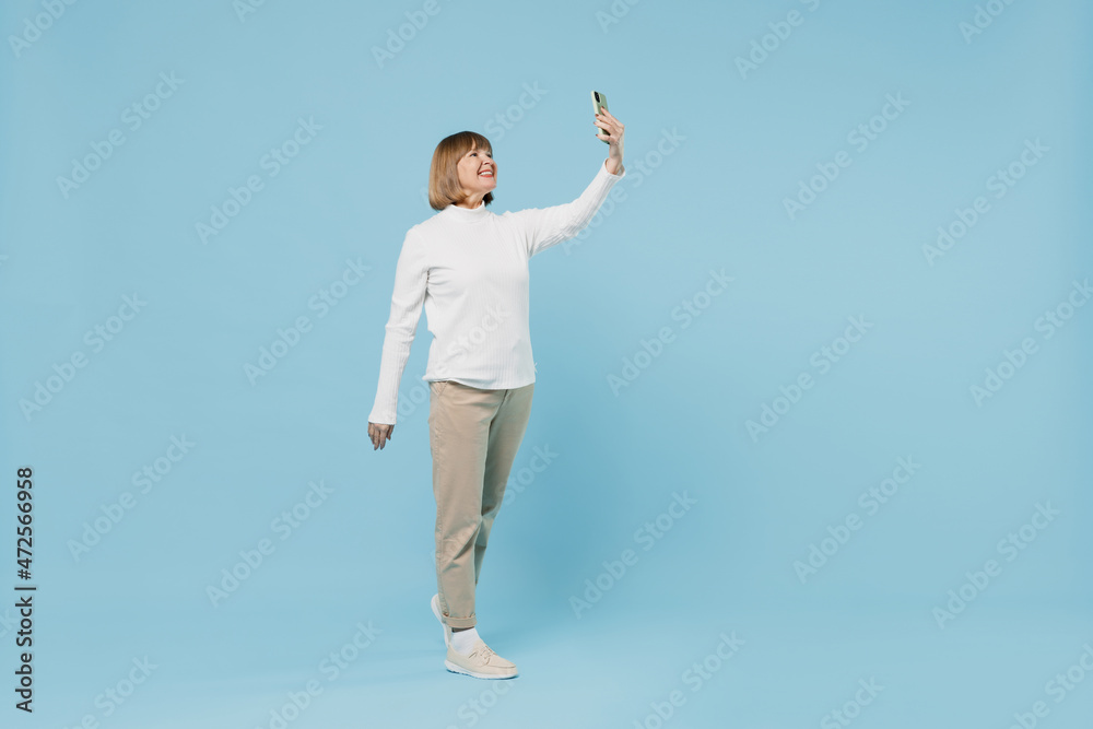 Full body elderly caucasian smiling woman 50s wearing white knitted sweater doing selfie shot on mobile cell phone isolated on plain blue color background studio portrait. People lifestyle concept Full body elderly caucasian smiling woman 50s wearing white knitted sweater doing selfie shot on mobile cell phone isolated on plain blue color background studio portrait. People lifestyle concept
