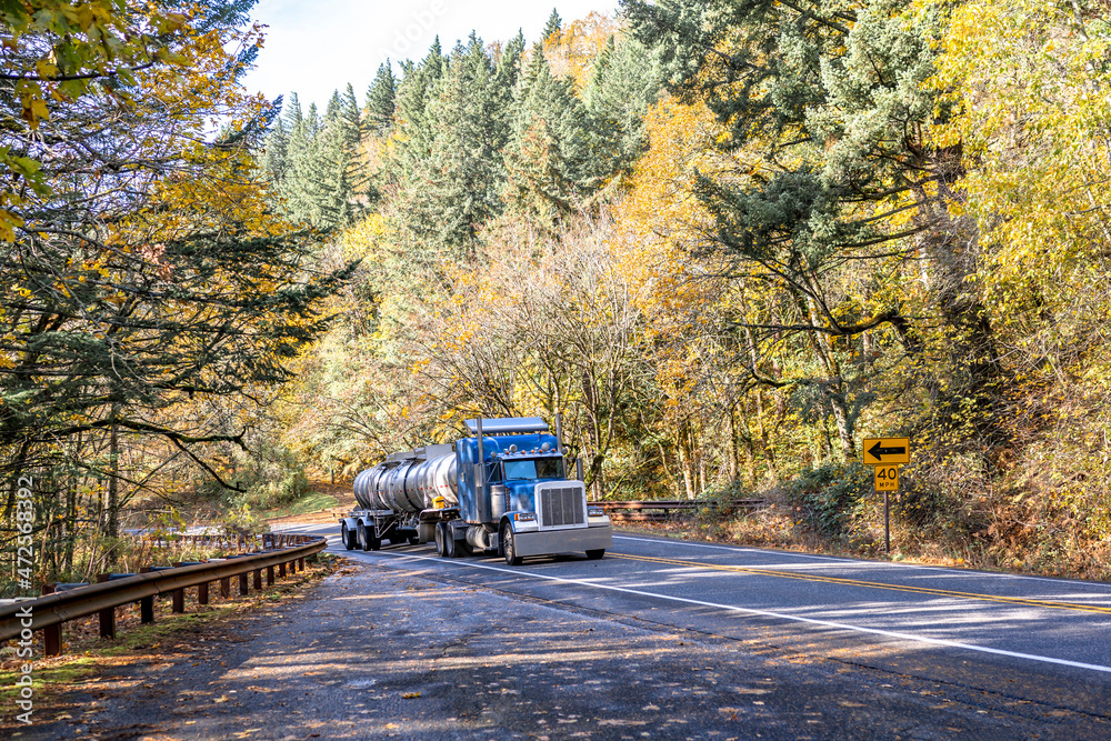 Classic powerful big rig blue semi truck transporting liquid cargo in armored tank semi trailer running on the winding mountain road with autumn forest