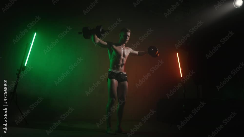 A sportsman is swings dumbbells to the sides. He is doing a shoulder exercise. See how his muscles are working. An orange and green light is shining on him. 4K A sportsman is swings dumbbells to the sides. He is doing a shoulder exercise. See how his muscles are working. An orange and green light is shining on him. 4K