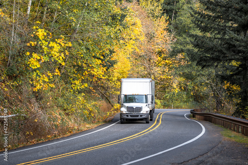 Medium class white rig semi truck with box trailer running on the winding road with autumn forest for the next delivery