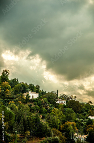 Cloudy scene with a hermitage among trees. Trees in autumn with clouds and warm light.