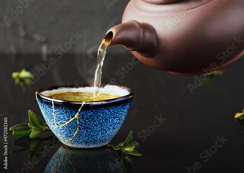 Hot green tea is poured from a ceramic teapot into a bowl. Selective focus on the blue cup. Steam will rise above the mug Reclaimed ceramic blue cup, second life of things, recycling or kintsugi photo