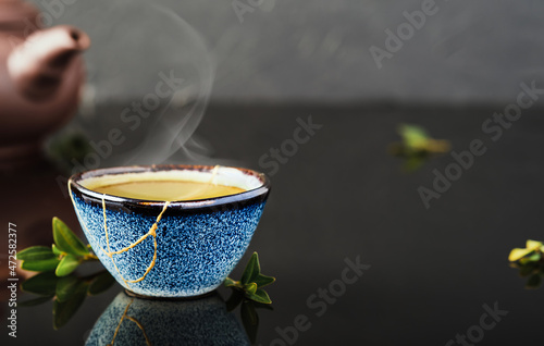 Bowl of green Japanese tea, tea leaves lie next the cup. Selective focus on the cup. Steam rises above the bowl. Reclaimed ceramic blue cup, second life of things, recycling or kintsugi photo