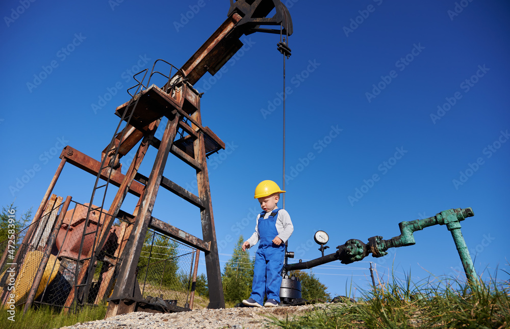 Bottom view of small young power engineer near oil rig. Boy in overalls against the blue sky, petroleum pipeline and pump jack for extracting natural fuel. Bottom view of small young power engineer near oil rig. Boy in overalls against the blue sky, petroleum pipeline and pump jack for extracting natural fuel.