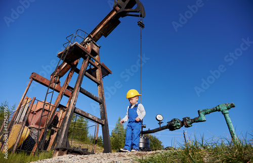 Fototapeta Naklejka Na Ścianę i Meble -  Bottom view of small young power engineer near oil rig. Boy in overalls against the blue sky, petroleum pipeline and pump jack for extracting natural fuel.