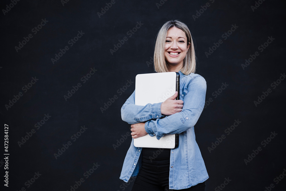 Contented female entrepreneur holding laptop in front of black wall Contented female entrepreneur holding laptop in front of black wall
