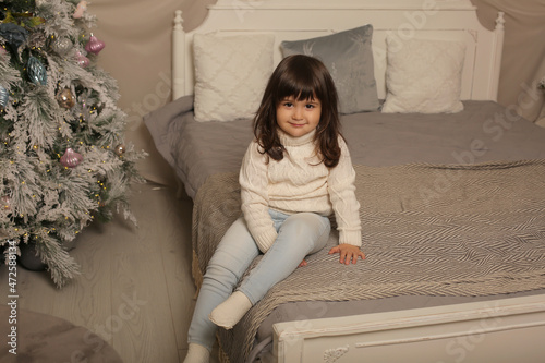 a beautiful little girl in a knitted sweater and jeans is sitting on the bed next to a Christmas tree