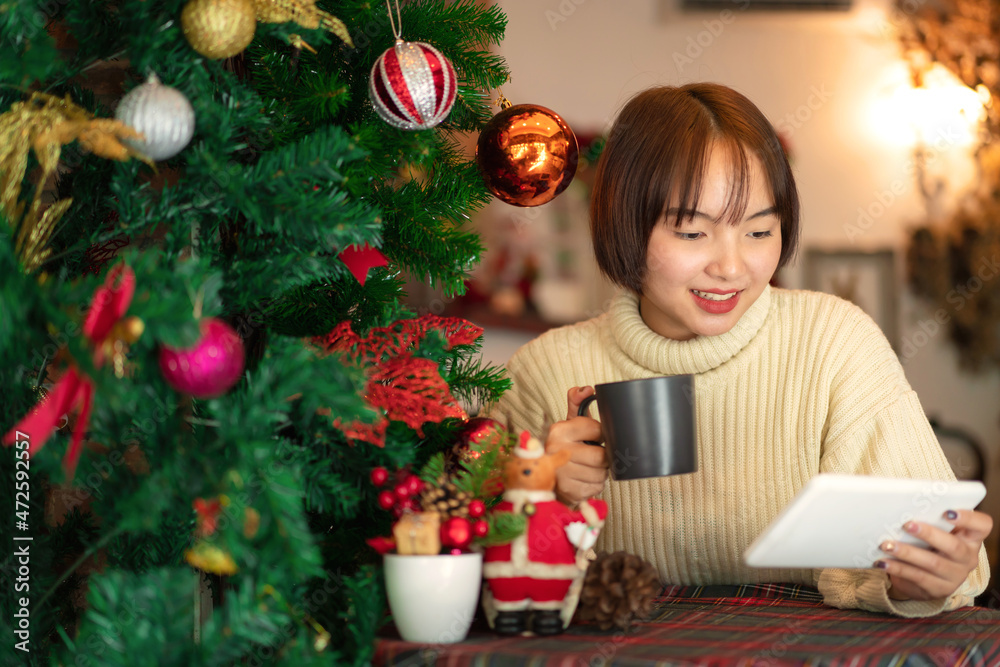 Beautiful woman in sweater sitting on chair to looking on tablet and drinking coffee cup near christmas tree Beautiful woman in sweater sitting on chair to looking on tablet and drinking coffee cup near christmas tree