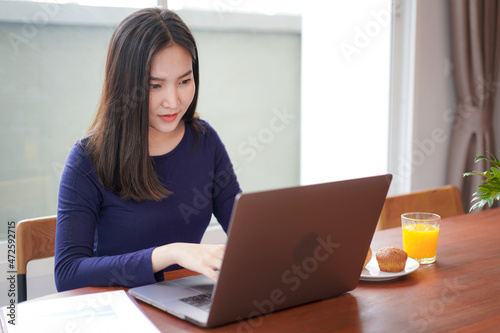 Young asian woman eating bread with a glass of orange juice while looking on laptop screen to typing