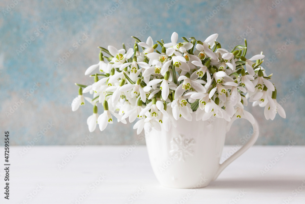 A bouquet of white spring flowers of snowdrops in a mug on a table against the background of a decorative wall, blur, selective focus. A bouquet of white spring flowers of snowdrops in a mug on a table against the background of a decorative wall, blur, selective focus.