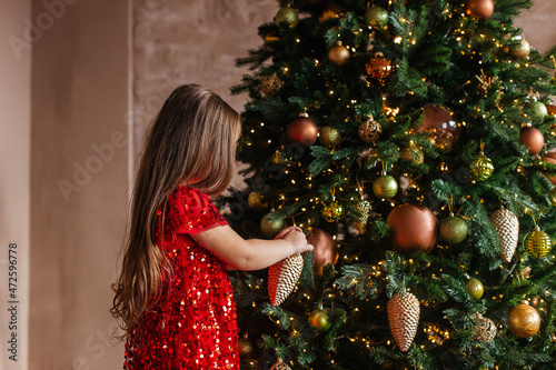 A little girl in a red dress with a Christmas tree toy in her hands stands at the Christmas tree, decorates it. Back view. Christmas Holidays. Christmas decor, atmosphere, winter background