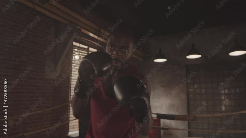 Handheld medium POV shot with slowmo of muscular Black man in boxing gloves looking at camera angrily while throwing boxing punches on ring Handheld medium POV shot with slowmo of muscular Black man in boxing gloves looking at camera angrily while throwing boxing punches on ring