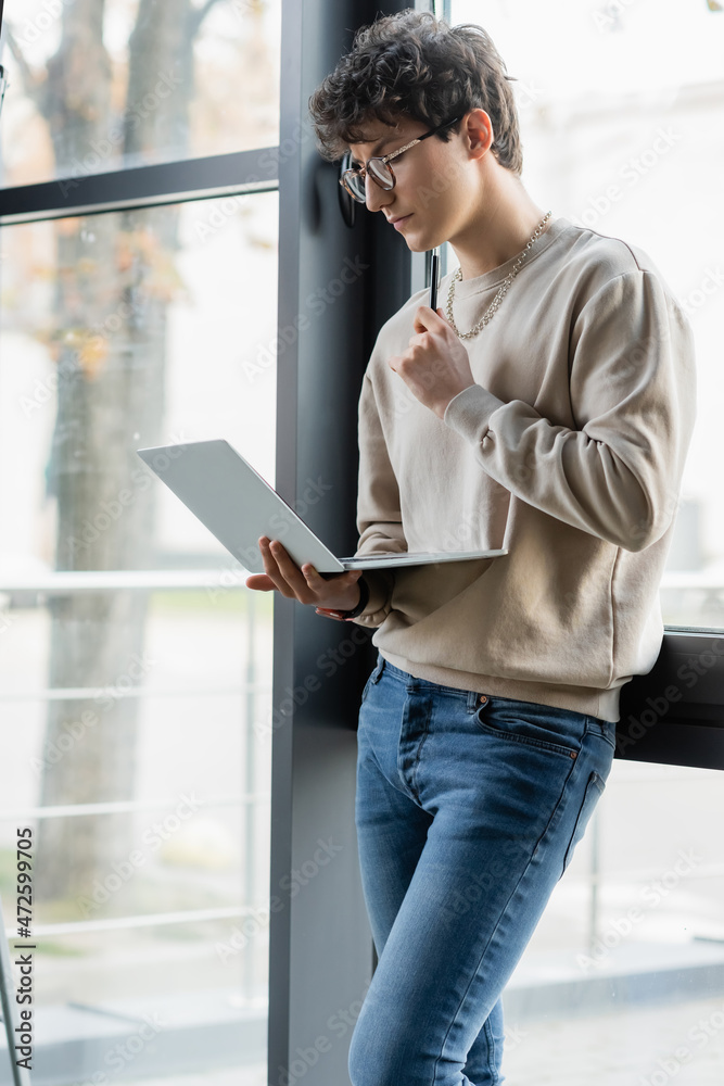 Fototapeta premium Young businessman holding pen and laptop near window in office.