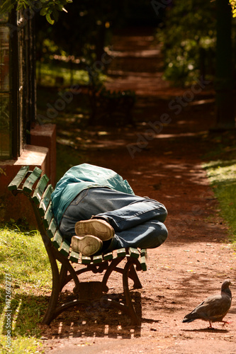 person sleeping on the street on a plaza bench, with a pigeon passing by, poverty, homelessness