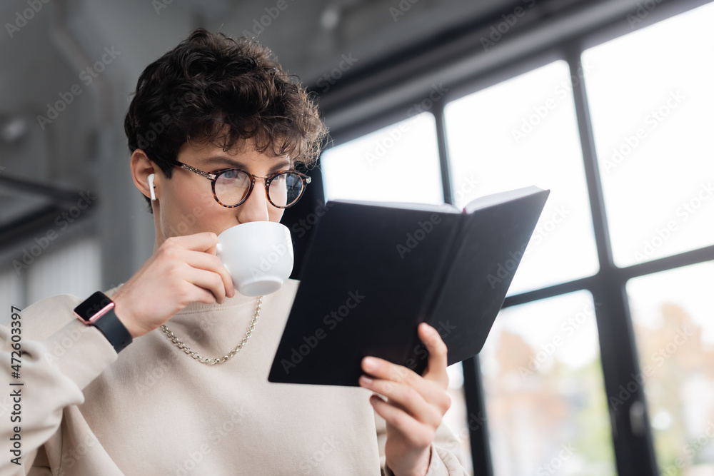 Young businessman in earphone drinking coffee and looking at notebook in office. Young businessman in earphone drinking coffee and looking at notebook in office.