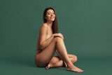 Young beautiful smiling girl in nude color underwear sitting on floor isolated over dark green studio background. Natural beauty concept.