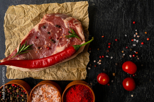 Raw piece of ribeye beef meat on craft paper on a wooden table stacked tomatoes garnished with hot peppers ,spice and himalayan salt. Fresh food. Everything for a barbecue.
