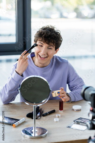 Smiling transgender person applying face powder near mirror, decorative cosmetics and digital camera.