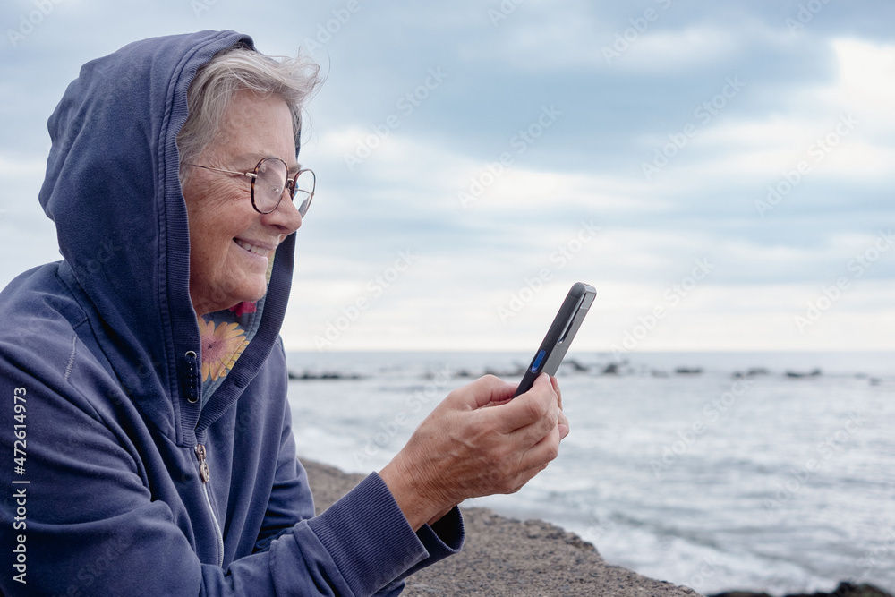 Smiling senior woman with hood on head standing at the sea in cloudy day holding smartphone messaging. 70s aged woman enjoying tech and social