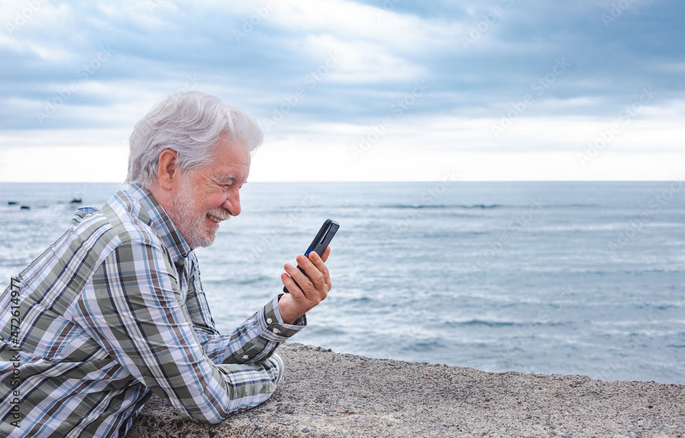 Attractive senior man smiling enjoying outdoors at sea using mobile phone. Cloudy sky and horizon over water Attractive senior man smiling enjoying outdoors at sea using mobile phone. Cloudy sky and horizon over water