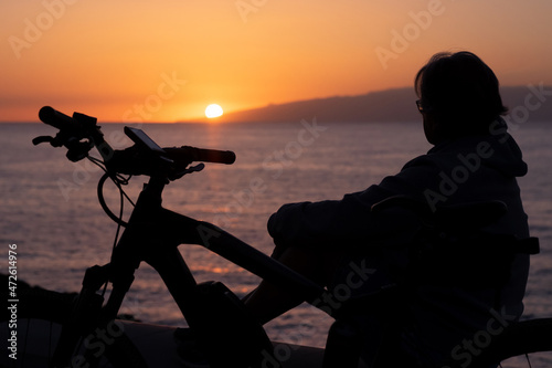 Silhouette of woman with electric bicycle sitting after cycling along the sea looking at sunset. Orange-black background