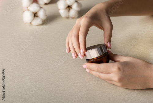 female hands open jar of cream for everyday care procedures against background of cotton flowers. Concept of beauty and health care.