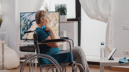 Aged person sitting in wheelchair using dumbbells to exercise while following online training lesson on tablet. Old woman with disability lifting weights and watching workout video.