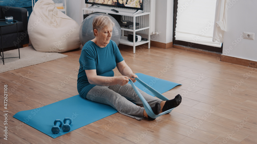 Old woman stretching arms and legs with resistance band, sitting on yoga mat.  Retired adult pulling elastic belt to do physical exercise and sport. Woman  training with fitness equipment Stock Photo