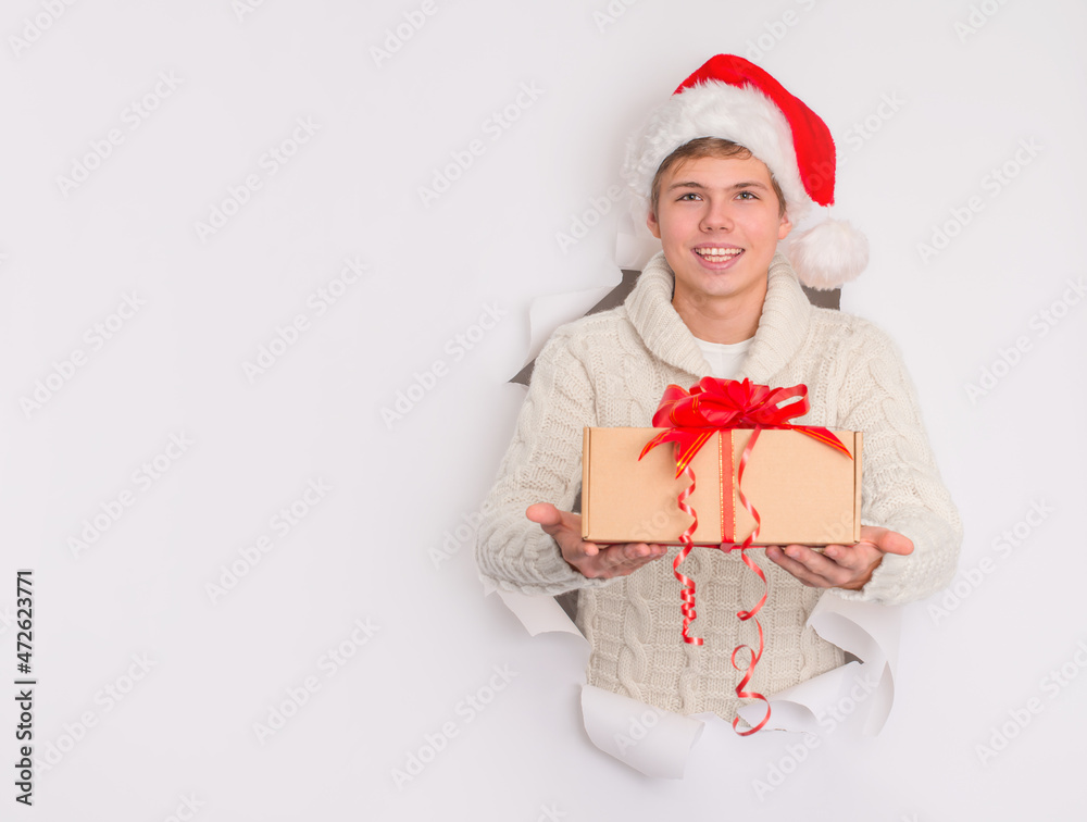 Happy teenager boy in Santa hat giving or delivering Christmas present gift box peeping out of the hole in white background. Happy teenager boy in Santa hat giving or delivering Christmas present gift box peeping out of the hole in white background.