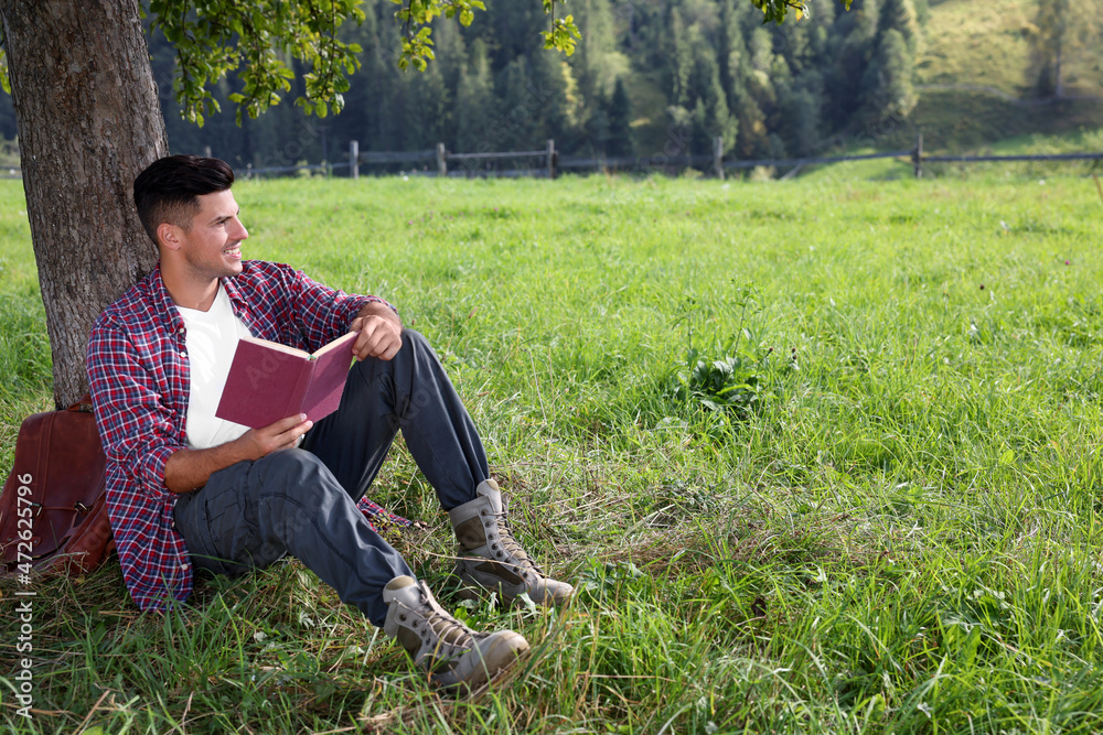 Handsome man reading book under tree on green meadow near forest Handsome man reading book under tree on green meadow near forest