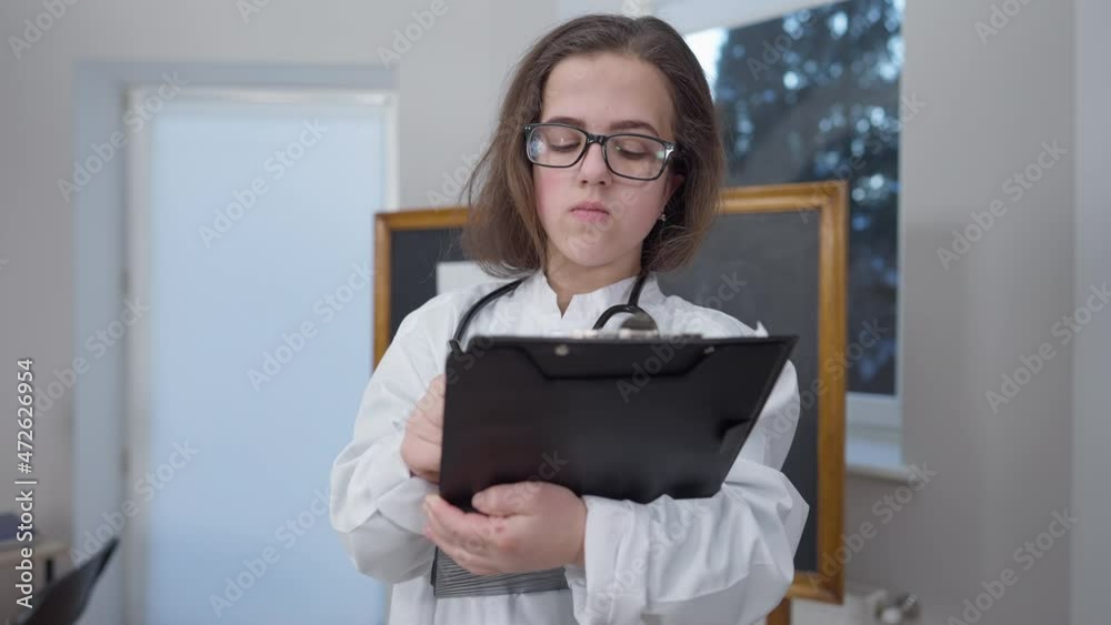 Medium shot concentrated female doctor in eyeglasses thinking writing with pen standing in hospital. Portrait of thoughtful Caucasian woman little person planning illness treatment indoors Medium shot concentrated female doctor in eyeglasses thinking writing with pen standing in hospital. Portrait of thoughtful Caucasian woman little person planning illness treatment indoors