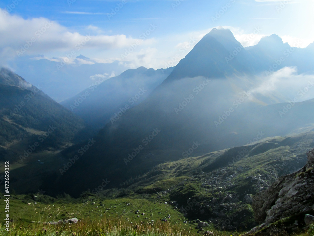 A stunning view on Alpine valley. Hiking trails in the high mountain. Green grass and bushes cover the slopes. Endless chains of mountains visible. Partially cloudy day. Spring in the mountains. A stunning view on Alpine valley. Hiking trails in the high mountain. Green grass and bushes cover the slopes. Endless chains of mountains visible. Partially cloudy day. Spring in the mountains.