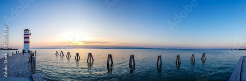 Fototapeta Naklejka Na Ścianę i Meble -  White and red striped lighthouse in Neusiedlersee, on Austrian-Hungarian border. Sun sets over the horizon in the clouds. Sky is orange, lake surface is soft and tender. Few clouds on the sky.
