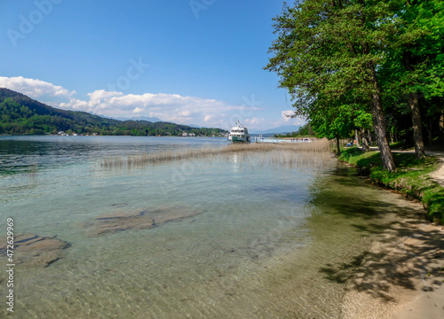 A boat parked on the shore of a lake. The water of the lake is crystal clear, the bottom is visible. In the back there are some mountains. Clear and bright day. Few trees on the side. photo