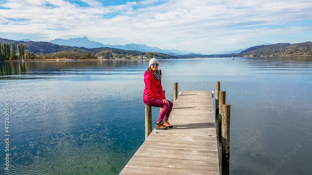 Girl wearing a pink jacket and beanie sits at the pillar of a promenade on the lake. Soft reflections of clouds in the lake. Clear but cloudy day. High mountains in the back. Calmness and relaxation