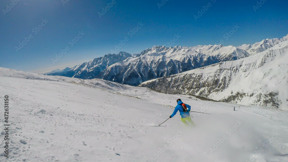 A skier going down the slope in Heiligenblut, Austria. Perfectly groomed slopes. High mountains surrounding the man wearing yellow trousers and blue jacket. Man wears helm for the protection.