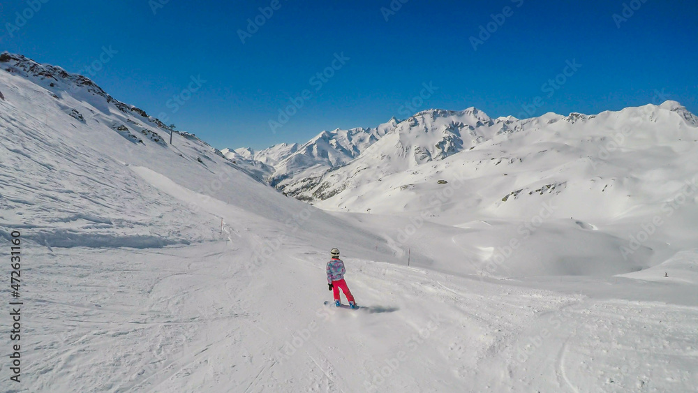 A snowboarder going down the slope in Heiligenblut, Austria. Perfectly groomed slopes. High mountains surrounding the girl, wearing pink trousers and colorful jacket. Girl wears helm for protection A snowboarder going down the slope in Heiligenblut, Austria. Perfectly groomed slopes. High mountains surrounding the girl, wearing pink trousers and colorful jacket. Girl wears helm for protection