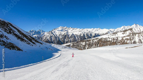 A snowboarder going down the slope in Heiligenblut, Austria. Perfectly groomed slopes. High mountains surrounding the man wearing yellow trousers and blue jacket. Girl wears helm for the protection.