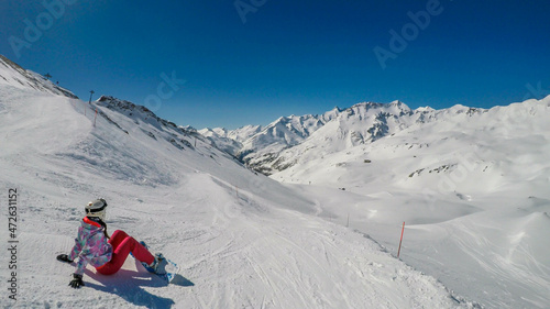 Snowboarder girl sitting on the slope admiring the tall Alps. Slopes are covered with powder snow. Perfect weather for a ride. High alpine snowboarding. Girl wears a helmet for protection.