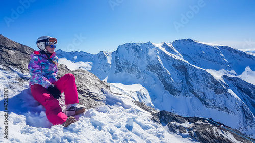 A snowboarder girl sitting on a slope admiring the tall Alps in front of her. Slopes are perfectly graveled. Perfect weather for a ride. High alpine snowboarding. Girl wears a helmet for protection.