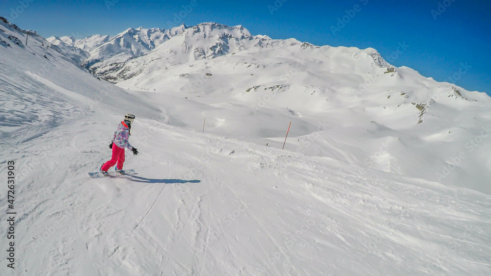 A snowboarder going down the slope in Heiligenblut, Austria. Perfectly groomed slopes. High mountains surrounding the girl, wearing pink trousers and colorful jacket. Girl wears helm for protection