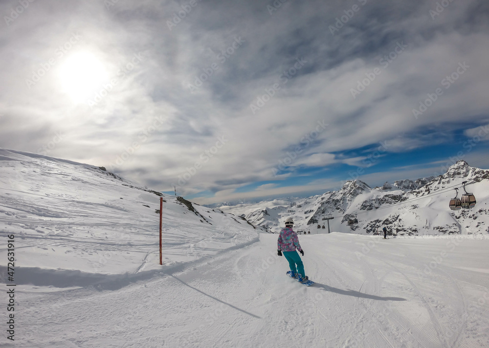 A snowboarder going down the slope in Moelltaler Gletscher, Austria. Perfectly groomed slopes. High mountains surrounding the girl wearing colourful snowboard outfit. Girl wears helm