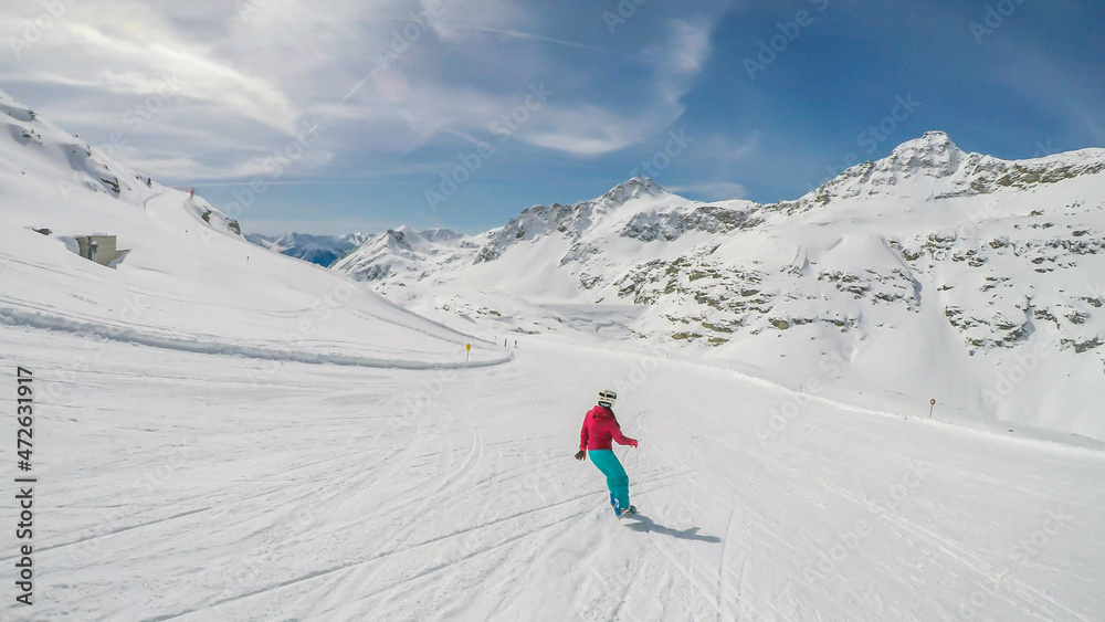 A snowboarder going down the slope in Moelltaler Gletscher, Austria. Perfectly groomed slopes. High mountains surrounding the girl wearing colourful snowboard outfit. Girl wears helm
