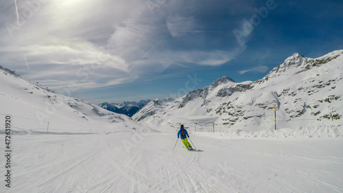 A skier going down the slope in Moelltaler Gletscher, Austria. Perfectly groomed slopes. High mountains surrounding the man wearing yellow trousers and blue jacket. Man wears helm for the protection.