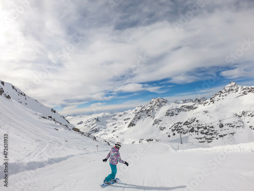 A snowboarder going down the slope in Moelltaler Gletscher, Austria. Perfectly groomed slopes. High mountains surrounding the girl wearing colourful snowboard outfit. Girl wears helmet