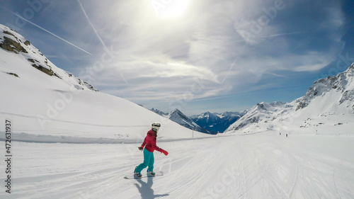 A snowboarder going down the slope in Moelltaler Gletscher, Austria. Perfectly groomed slopes. High mountains surrounding the girl wearing colourful snowboard outfit. Girl wears helmet