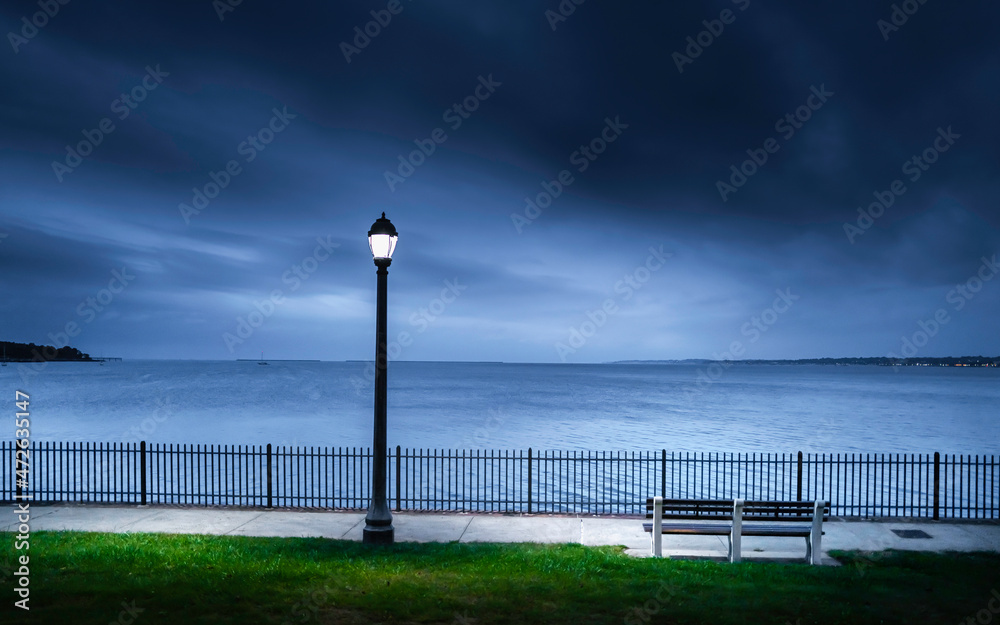 Dramatic twilight seascape with a street lamp, bench, a metal fence over the green in the beach park. Tranquil moody landscape over the boardwalk in New Haven Harbor, Connecticut.