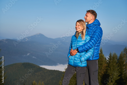 Handsome man hugging his smiling girlfriend from behind and they together standing and watching the surrounding nature against the mountain hills on sunny day.